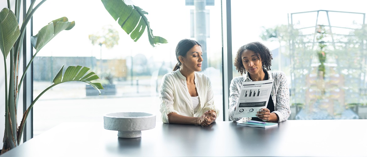 Man sat at table, woman in suit stood at printer in office, chairs, tables, plants, people in the background